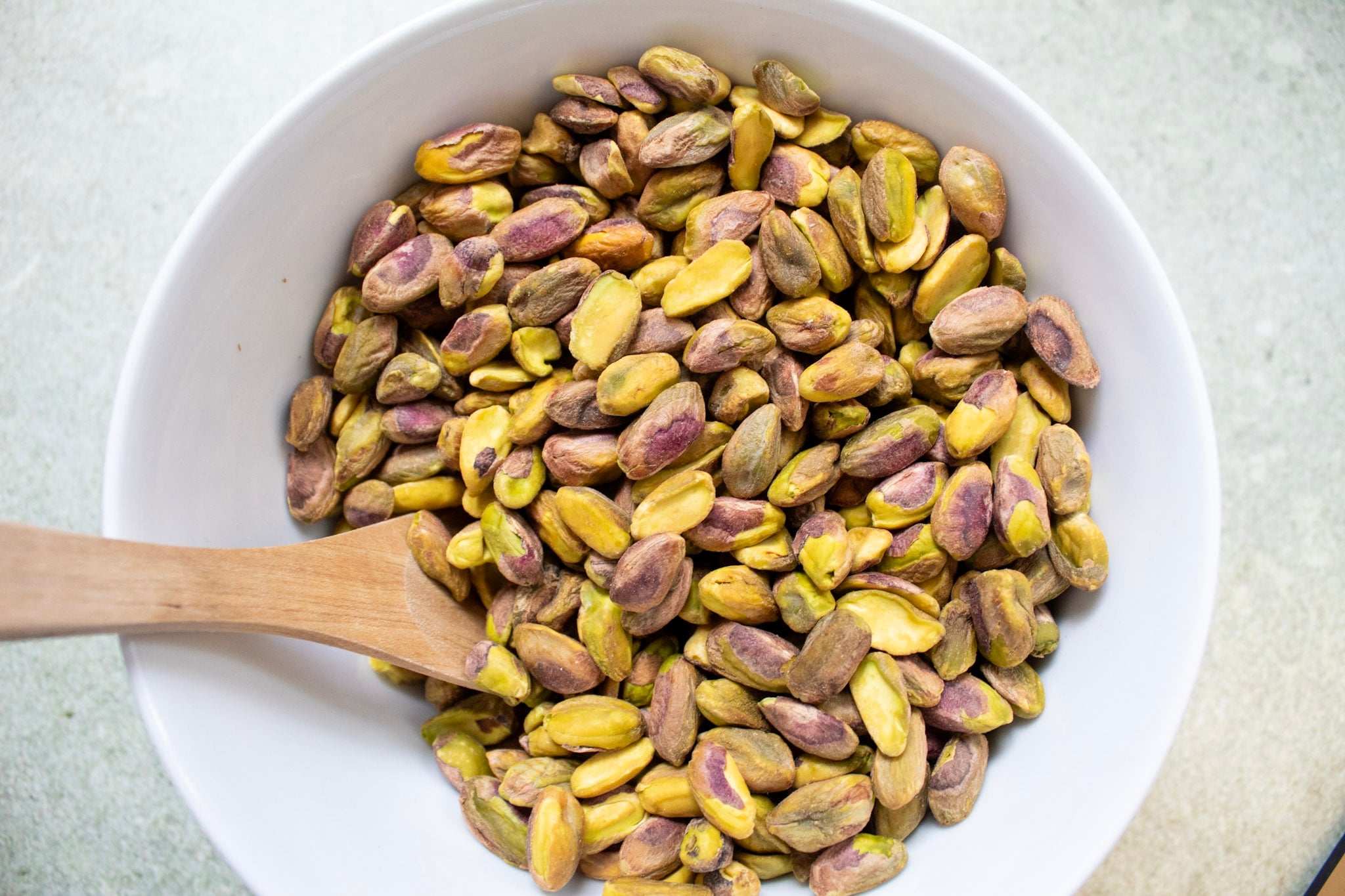 A photo of a bowl of whole pistachio nuts with wooden spoon.