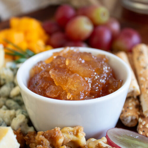 a bowl of apple cider jam on a cheese board surrounded by cheese and crostini.