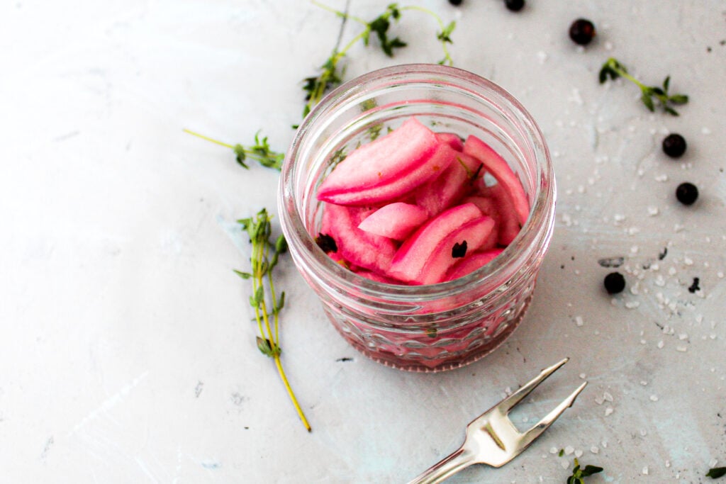 pickled red onion in glass jar with thyme herb and juniper berries. 